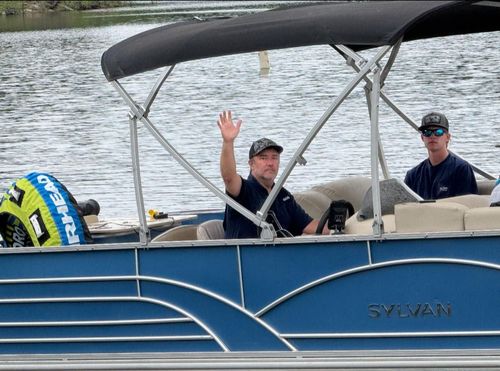 Two people in hats on a blue Sylvan pontoon boat, one waving to the camera while on the water.