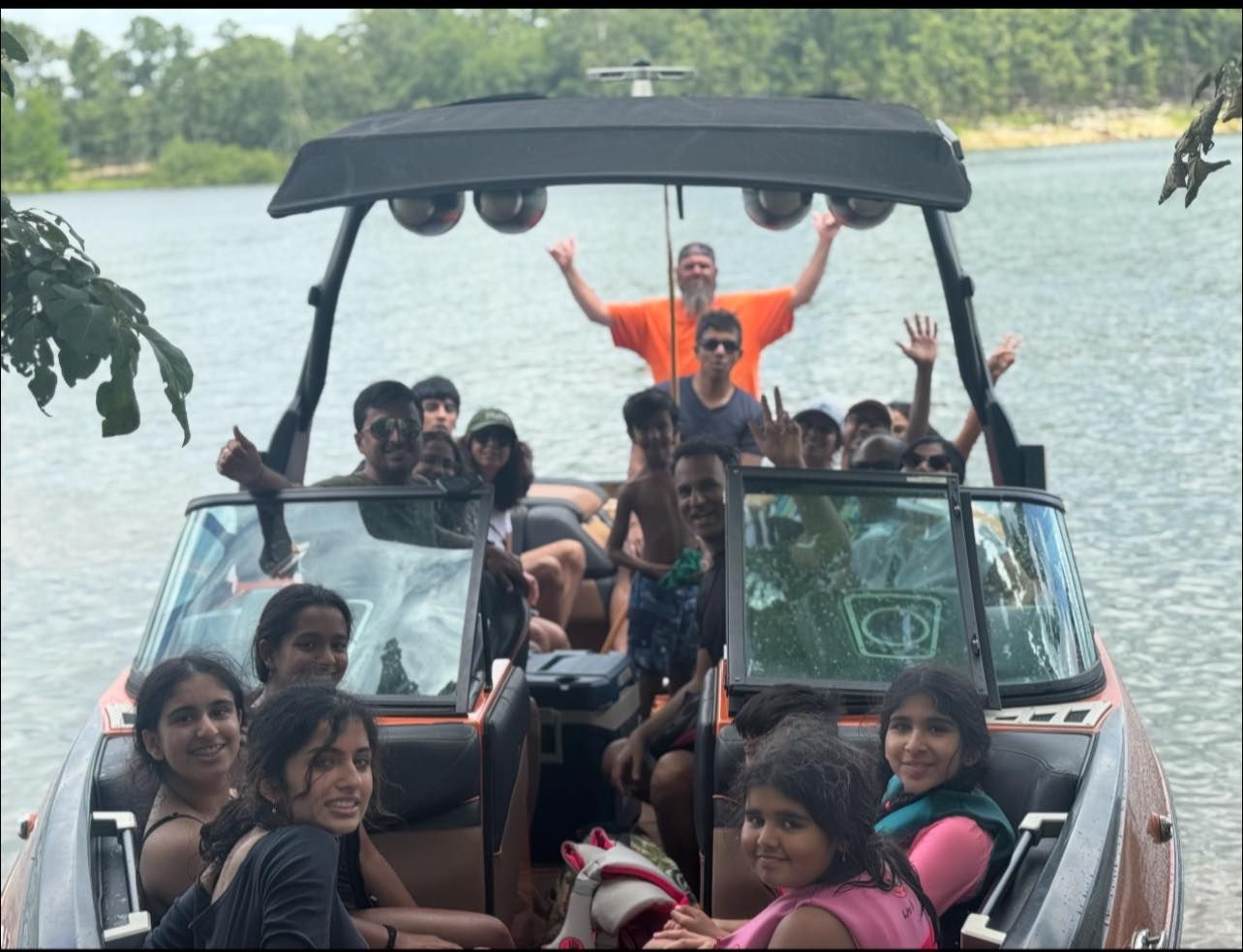 A group of people smiles and waves from a motorboat on a lake, with one person standing on the deck in an orange shirt.