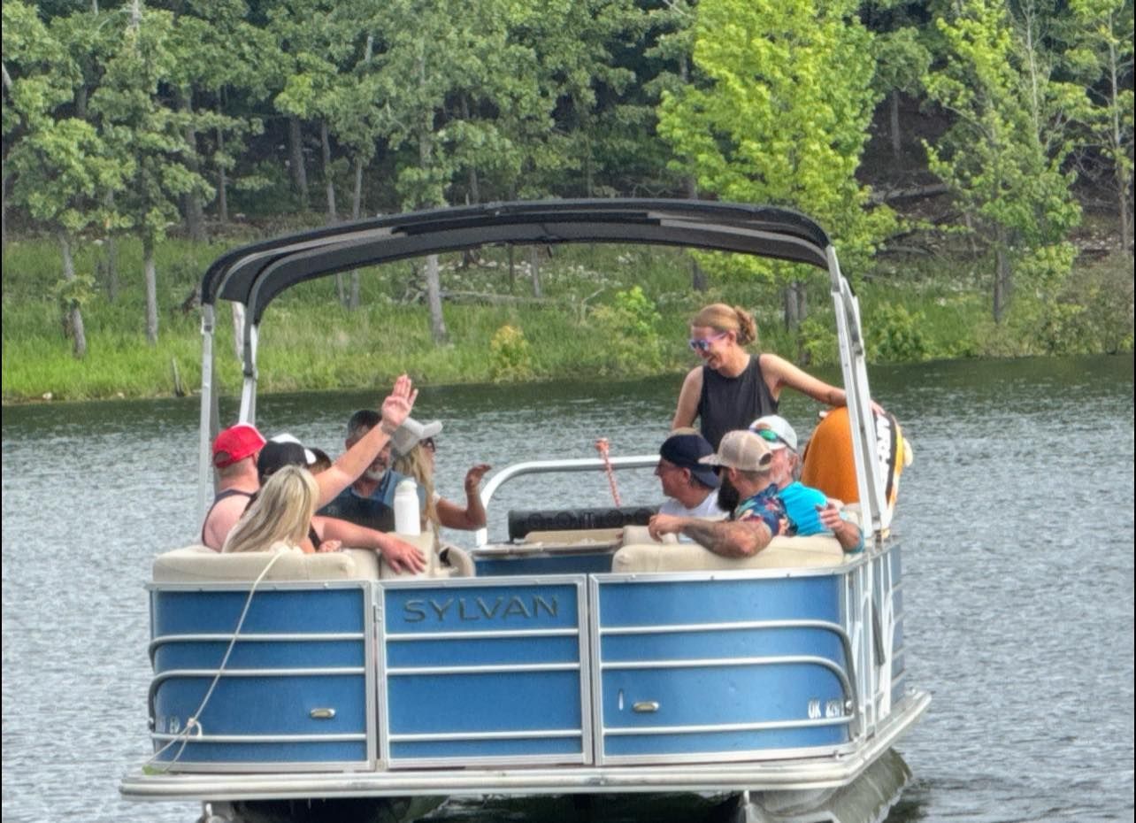 A group of people enjoys a sunny day on a blue pontoon boat moving across a lake with a wooded shoreline in the background.