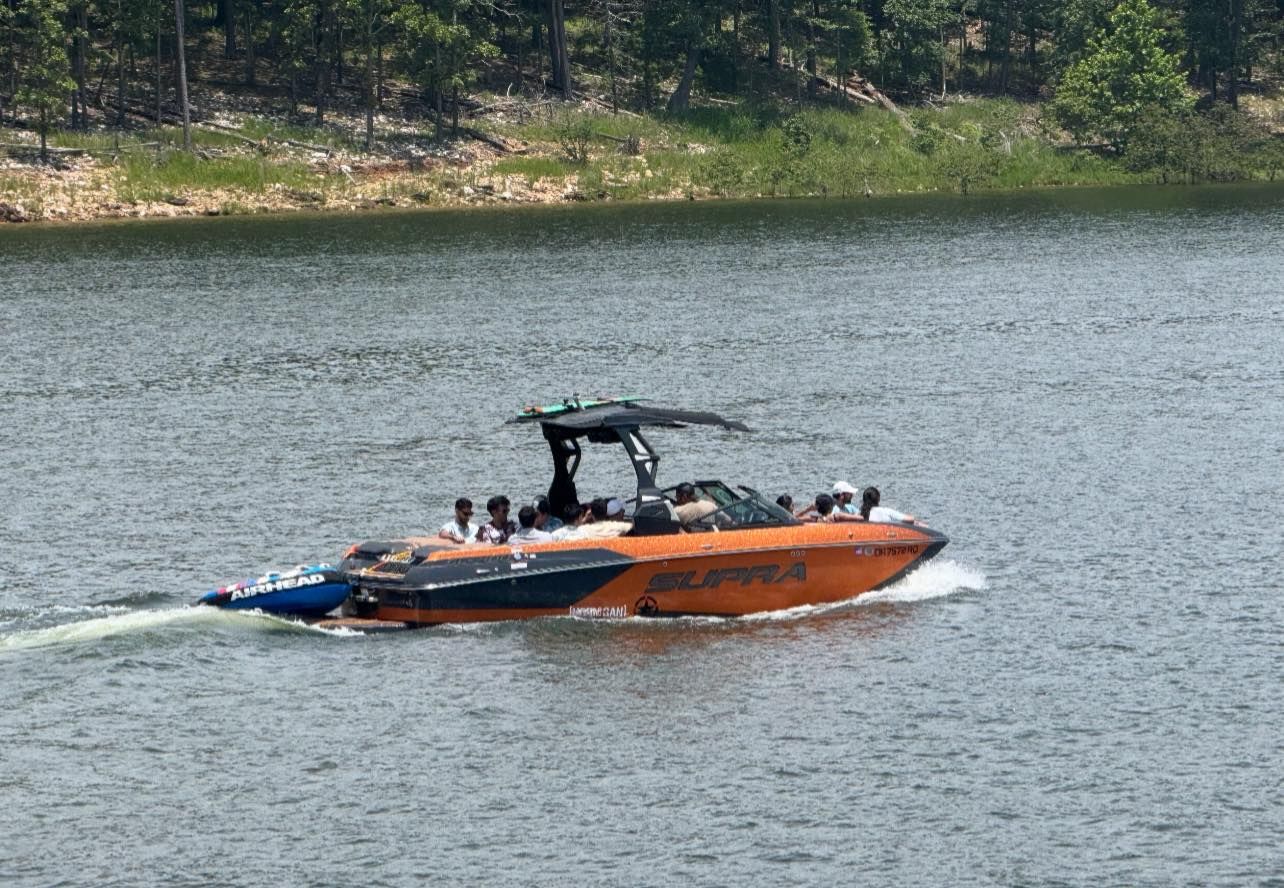 An orange and black Supra wake boat carrying a group of people moves across a lake near a tree-lined shore.