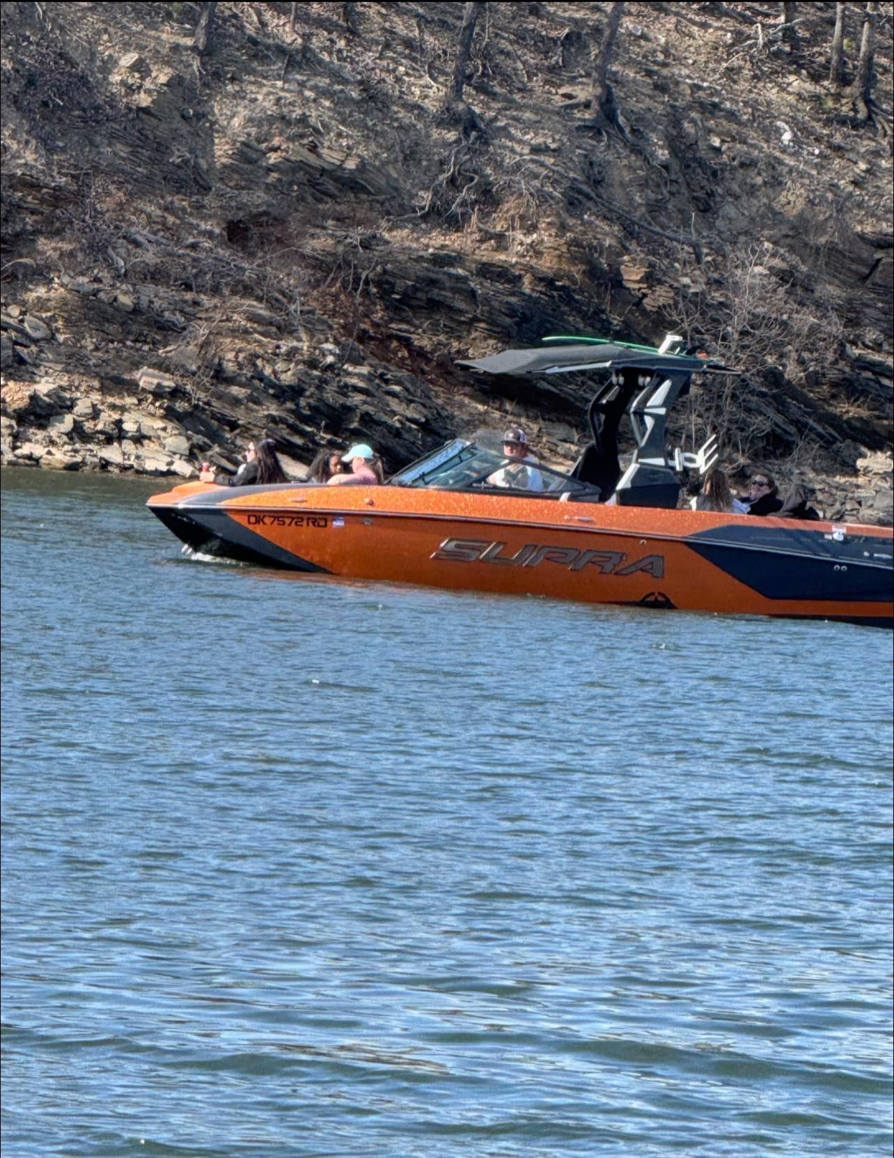 A bright orange and dark grey speedboat cruises on a lake in front of a steep, rocky, brush-covered shoreline.