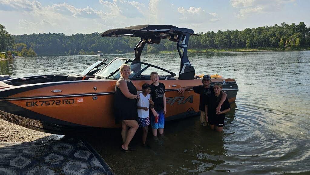 A group of five people stand in shallow water beside an orange motorboat on a lake shore with trees in the background.