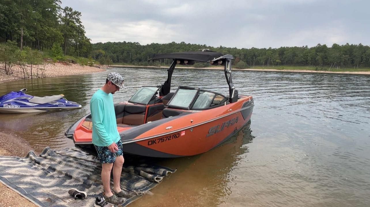 A person in a teal shirt standing next to an orange and black boat beached on a sandy shore by a lake.