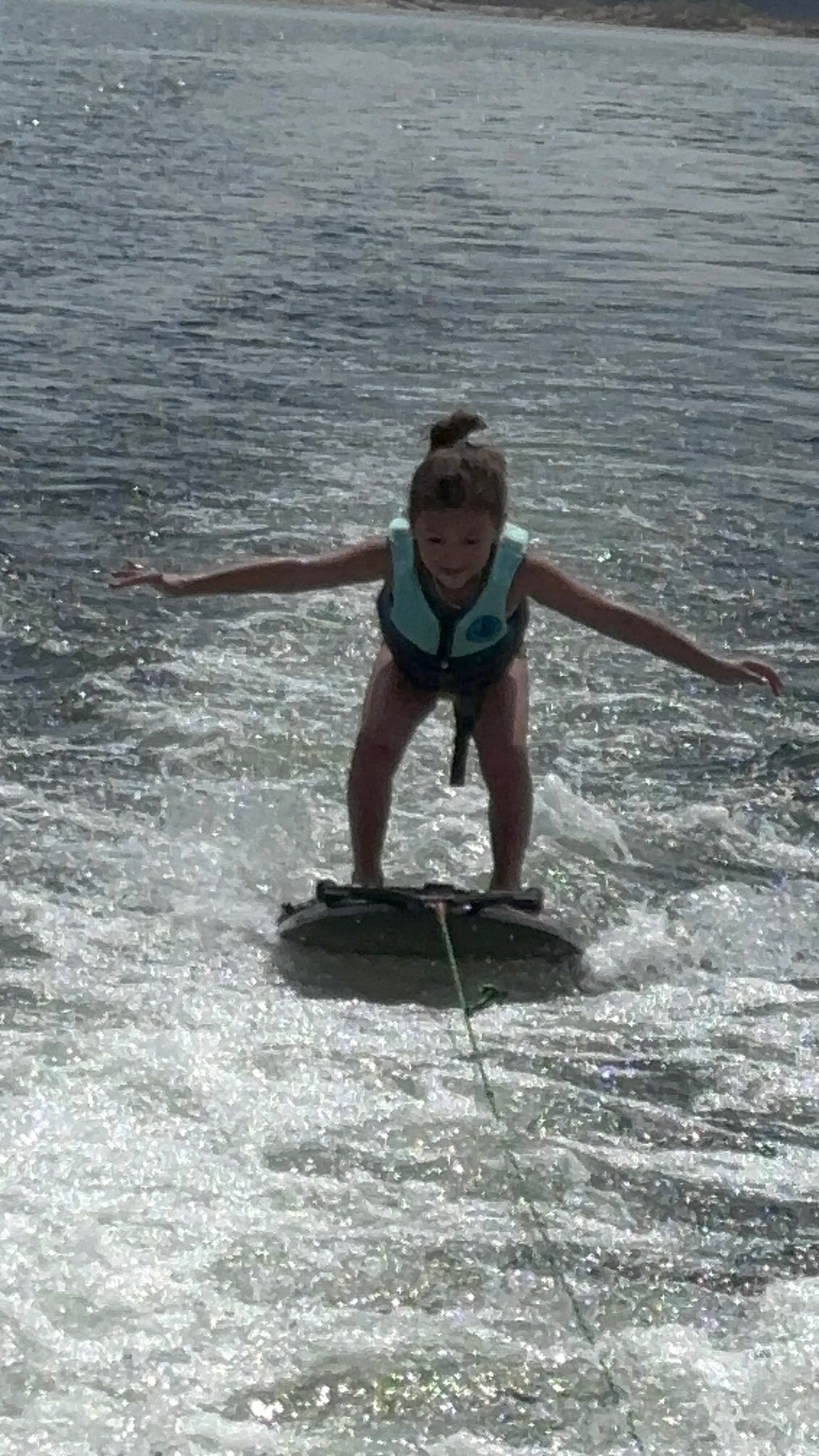 A person wearing a life vest kneels on a wakeboard, balanced and smiling, while gliding over water in a boat's wake.