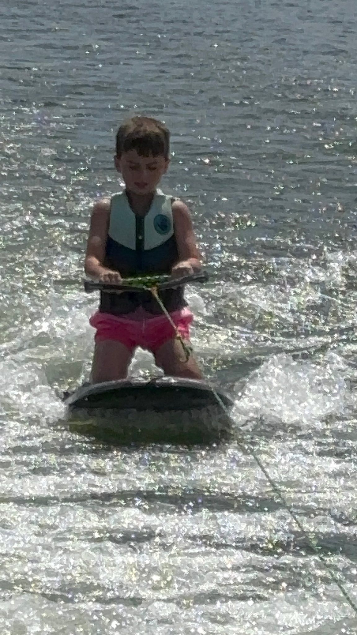 A person wearing a life vest and pink shorts kneels on a wakeboard, being towed across the water on a sunny day.