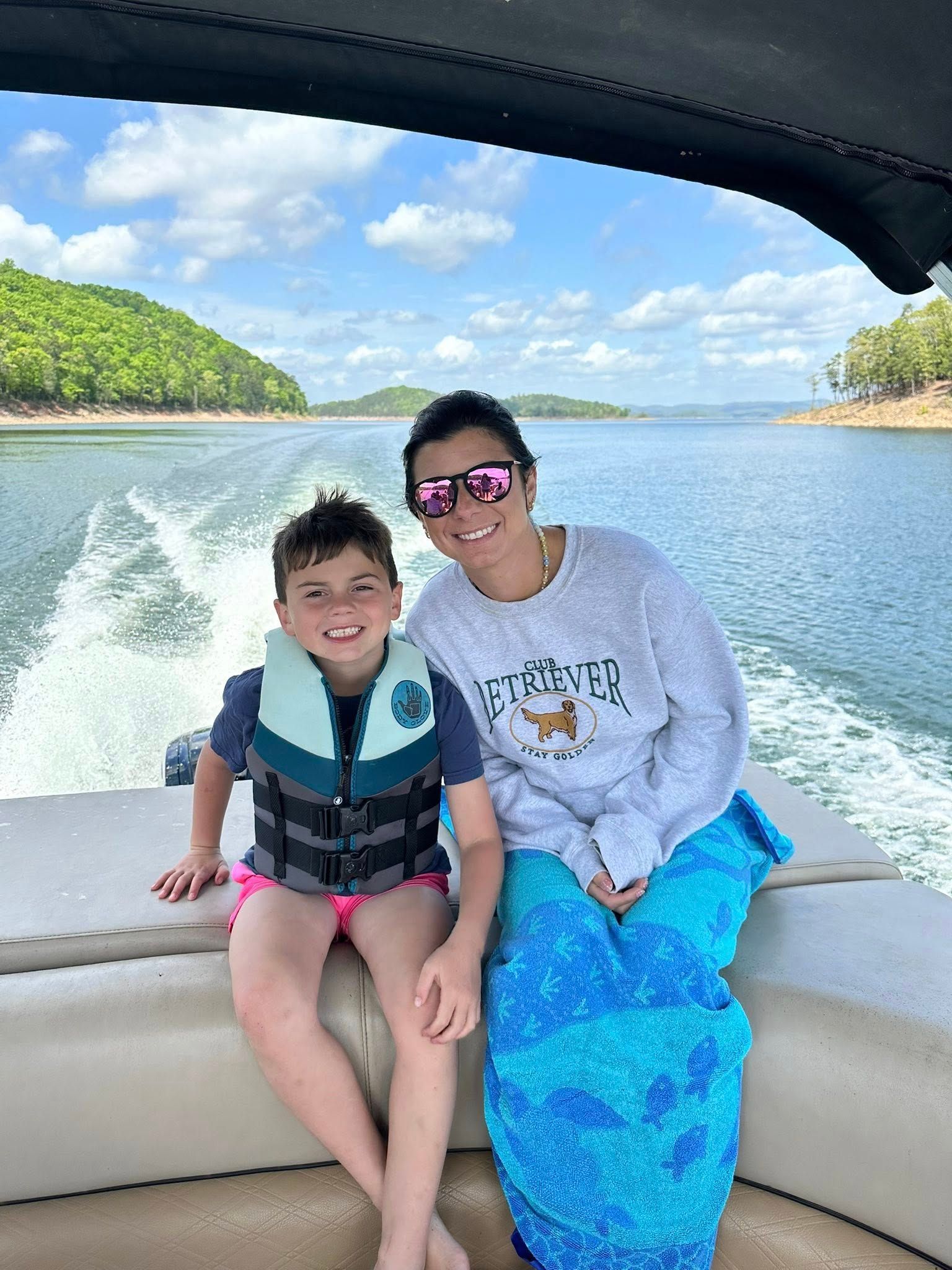A person and child in a life vest sit on a boat on a sunny lake, smiling at the camera with a boat wake behind them.