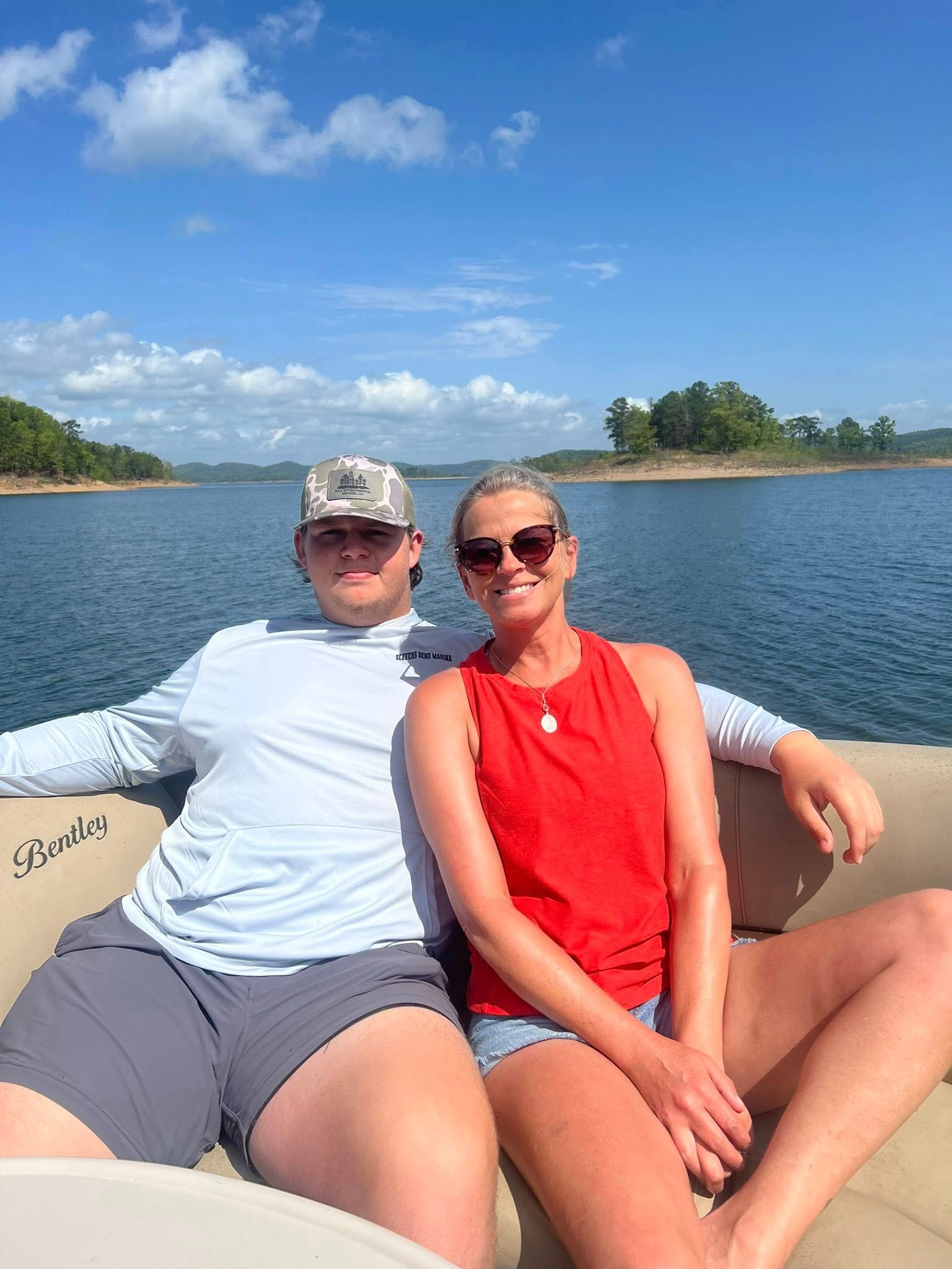 A person in a light shirt and cap and another person in a red tank top sitting together on a boat on a sunny lake.