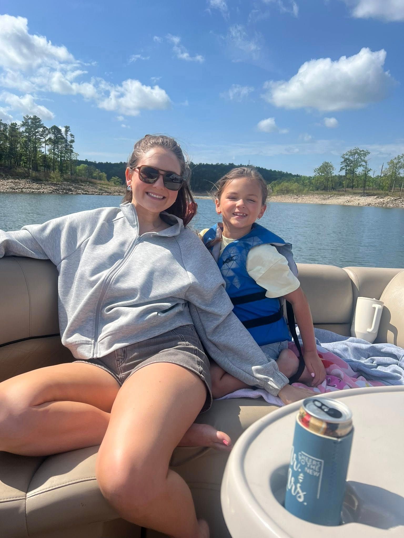 A person in a gray hoodie and shorts and a child in a blue life vest sit together on a boat on a sunny day.