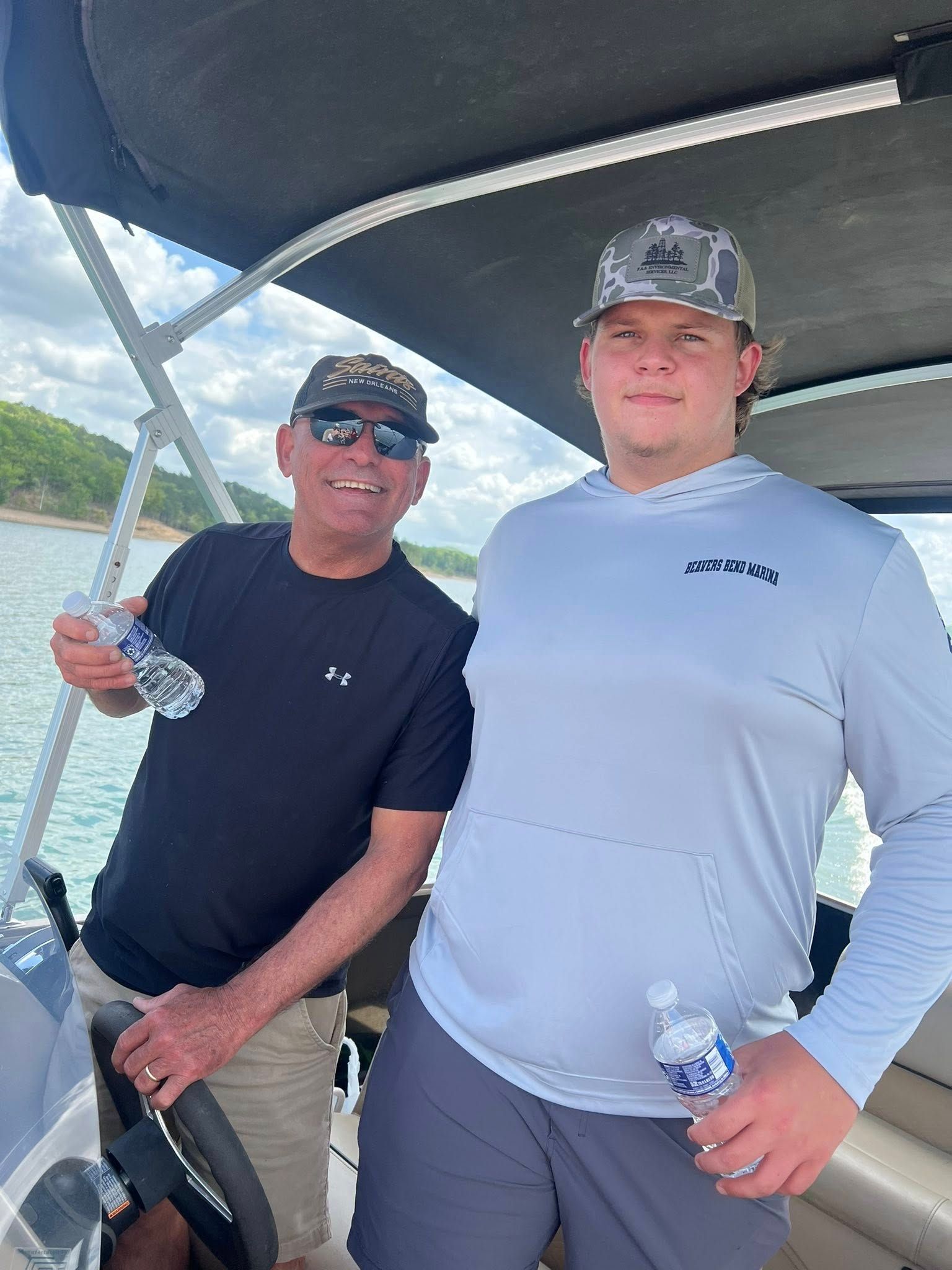 Two people wearing caps stand on a boat on the water, holding water bottles and smiling toward the camera.