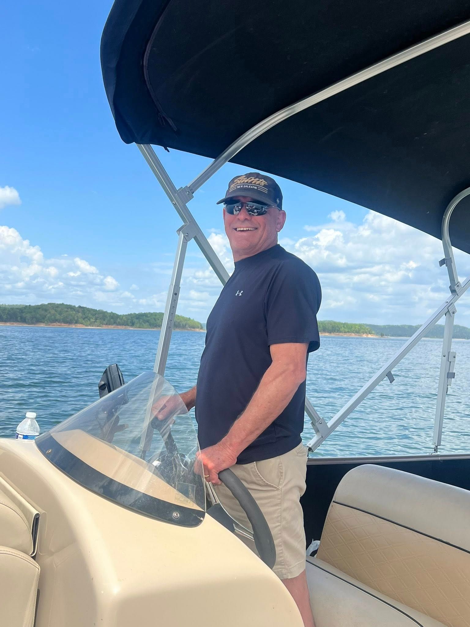 A person wearing sunglasses and a cap steering a pontoon boat on a sunny lake.