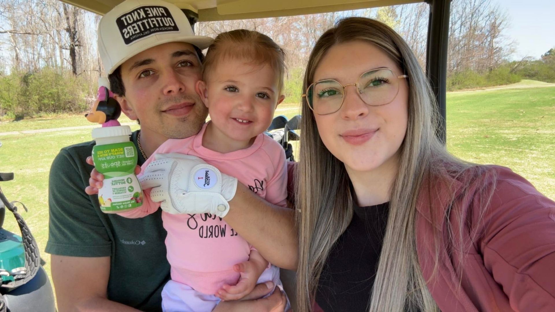 A man, woman, and small child smiling together in a golf cart on a sunny day.