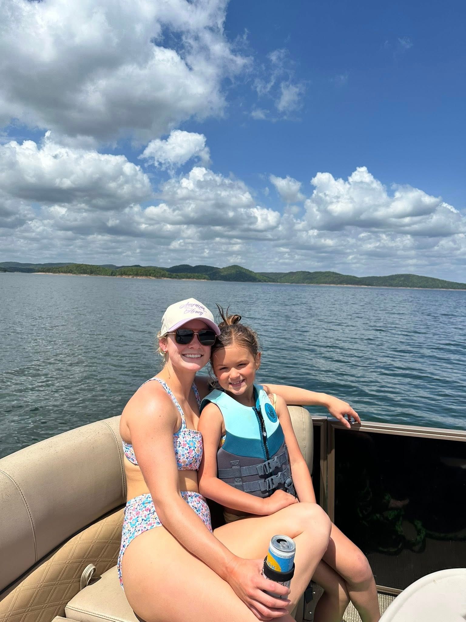A person wearing a floral swimsuit and hat sits on a boat with a child wearing a life vest on a sunny day at a lake.