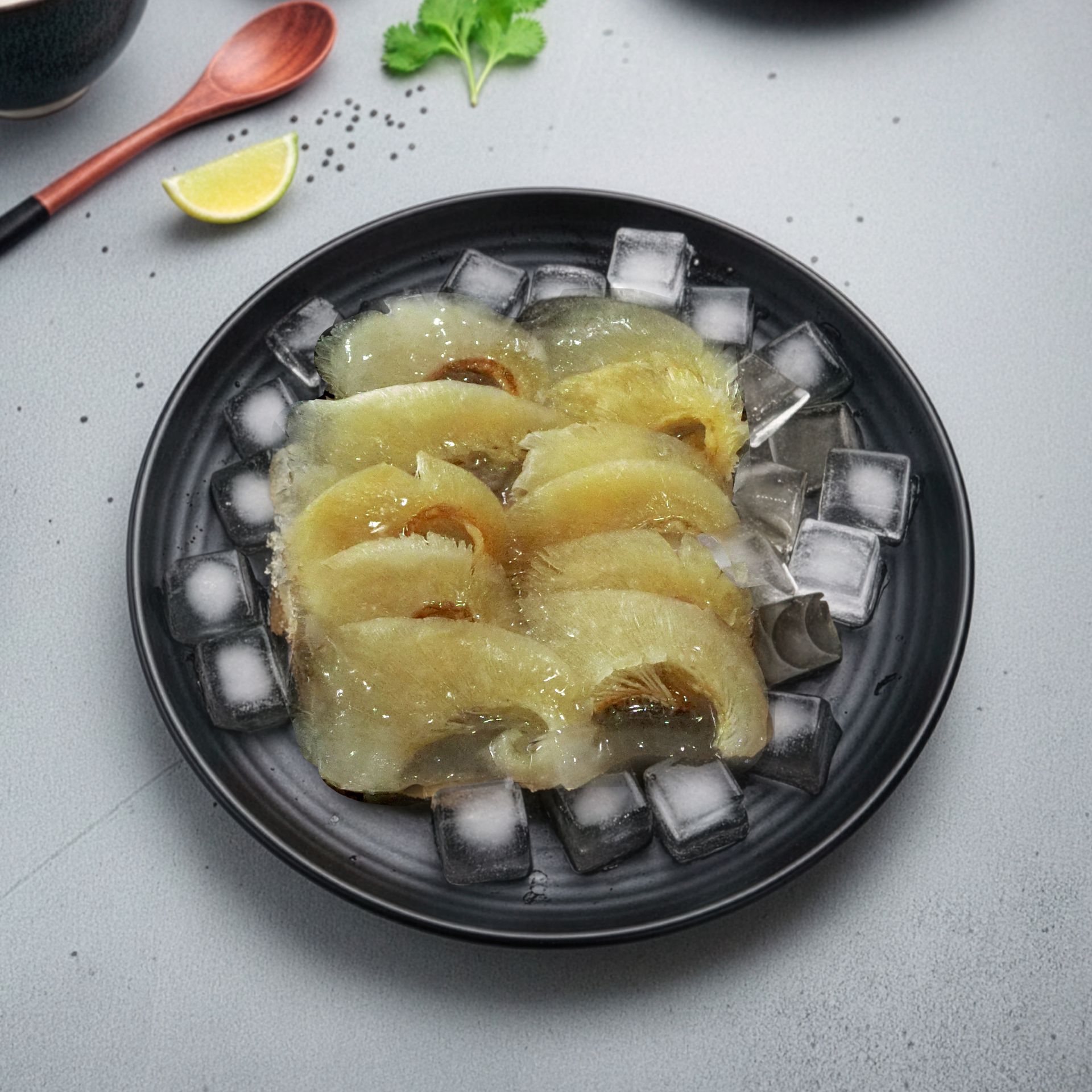 切片的金黃色食物盛放在黑色盤子裡,旁邊放著冰塊。旁邊還有木匙、檸檬和香草。