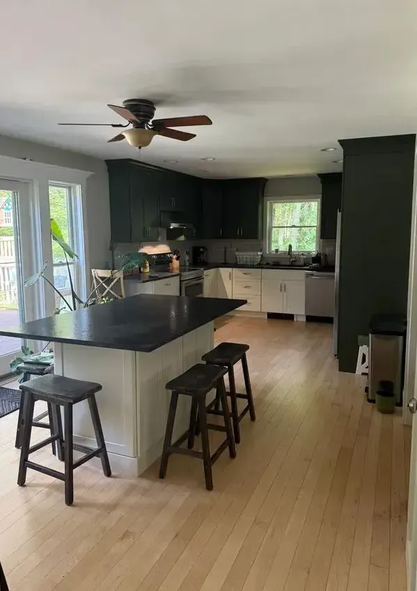 Bright kitchen with black island, bar stools, white cabinets, and a ceiling fan opening to a living area