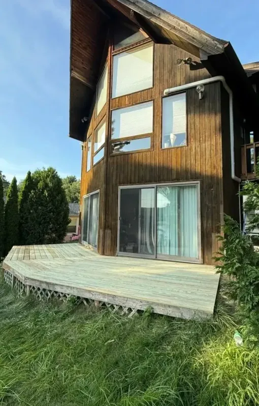 Wooden house with curved deck and large windows in a grassy yard under a blue sky
