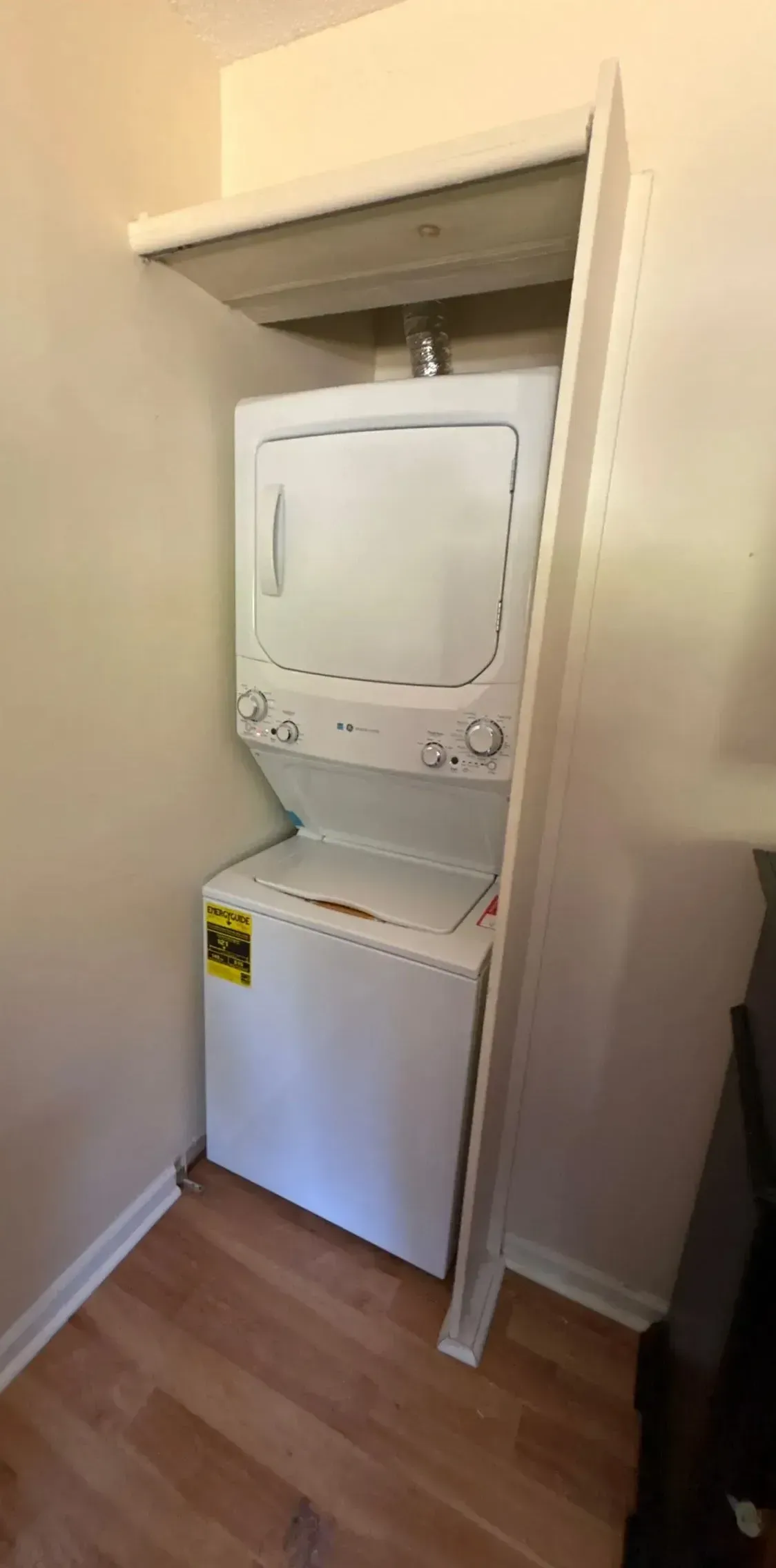 Stacked washer and dryer in a narrow closet with white walls and wood floor.