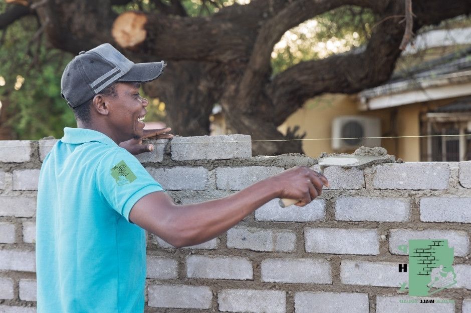 A block wall repair company worker in a gray cap and turquoise shirt carefully positions concrete blocks while building a residential wall. The smiling mason is creating a sturdy structure at this residential project near W Medlock Dr, and N 180th Ln in Citrus Park, AZ.