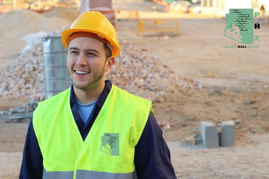 A smiling block wall repair company worker wearing an orange hard hat and bright yellow safety vest stands confidently at a commercial construction site. Behind him are piles of gravel and concrete blocks ready for the masonry project near N 9th St and Speer Trail, New River, AZ.
