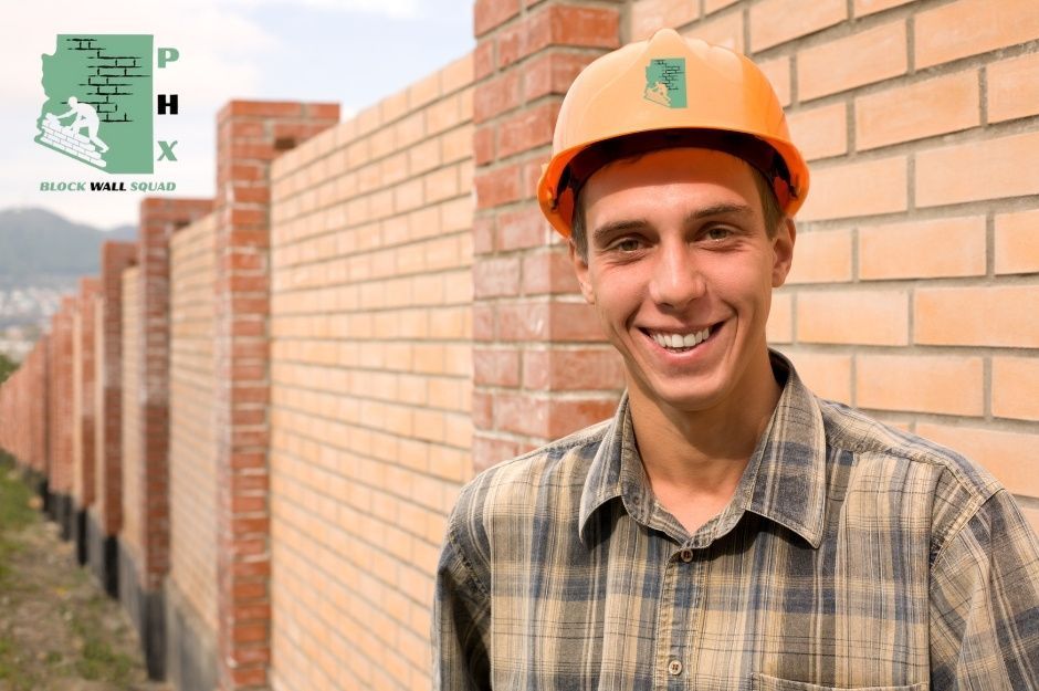Block wall repair company worker in an orange hard hat and plaid shirt smiling in front of completed brick and block wall construction. A professional contractor stands beside the tall commercial masonry wall with red brick pillars and tan block sections near W Crawfordsville Dr, and N Blakely St in Surprise, AZ.