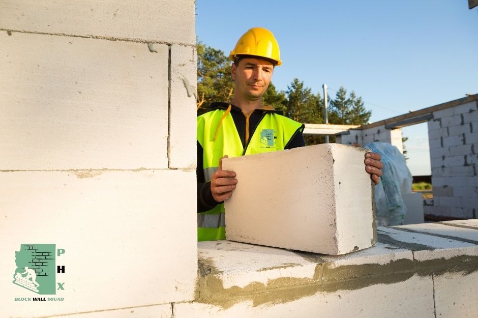 A professional block wall repair contractor in yellow hard hat and high-visibility vest installs concrete blocks for residential construction. He carefully positions blocks building the wall structure under clear blue skies near E Vía Los Caballos, and E Vía Del Cielo in Paradise Valley, AZ.