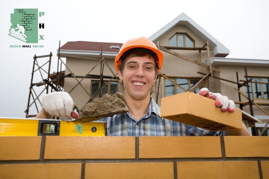 A block wall repair contractor wearing an orange hard hat and plaid shirt stands behind a row of concrete blocks, with a yellow spirit level tool. Construction scaffolding and a house under residential construction are visible in the background near N Paradise Found Trail, and N Pretty Penny Path in Prescott Valley, AZ.