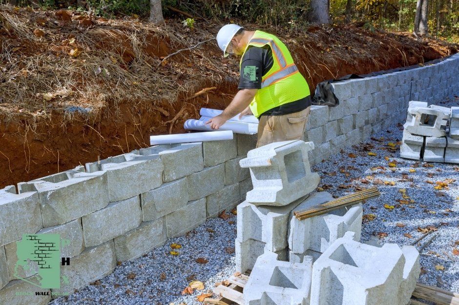 An expert block wall repair specialist in a bright yellow safety vest and white hard is reading a blue print for a commercial retaining wall project. Loose gravel and concrete blocks are visible on the ground around the work area. He is working near E Eagle Rock Dr, and N Boulder Dr in Fountain Hills, AZ.