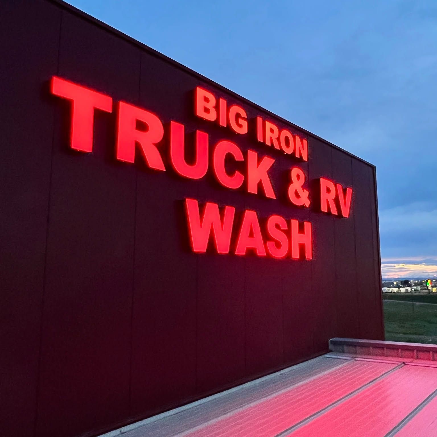 Red neon “BIG IRON TRUCK & RV WASH” sign on a dark building at dusk