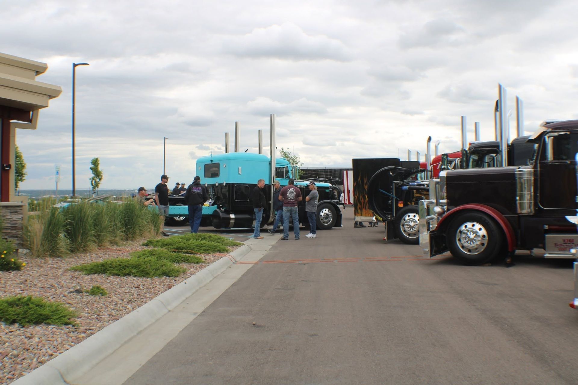 Truck gathering on a roadside, with parked black semis, a blue truck, and people under a cloudy sky.