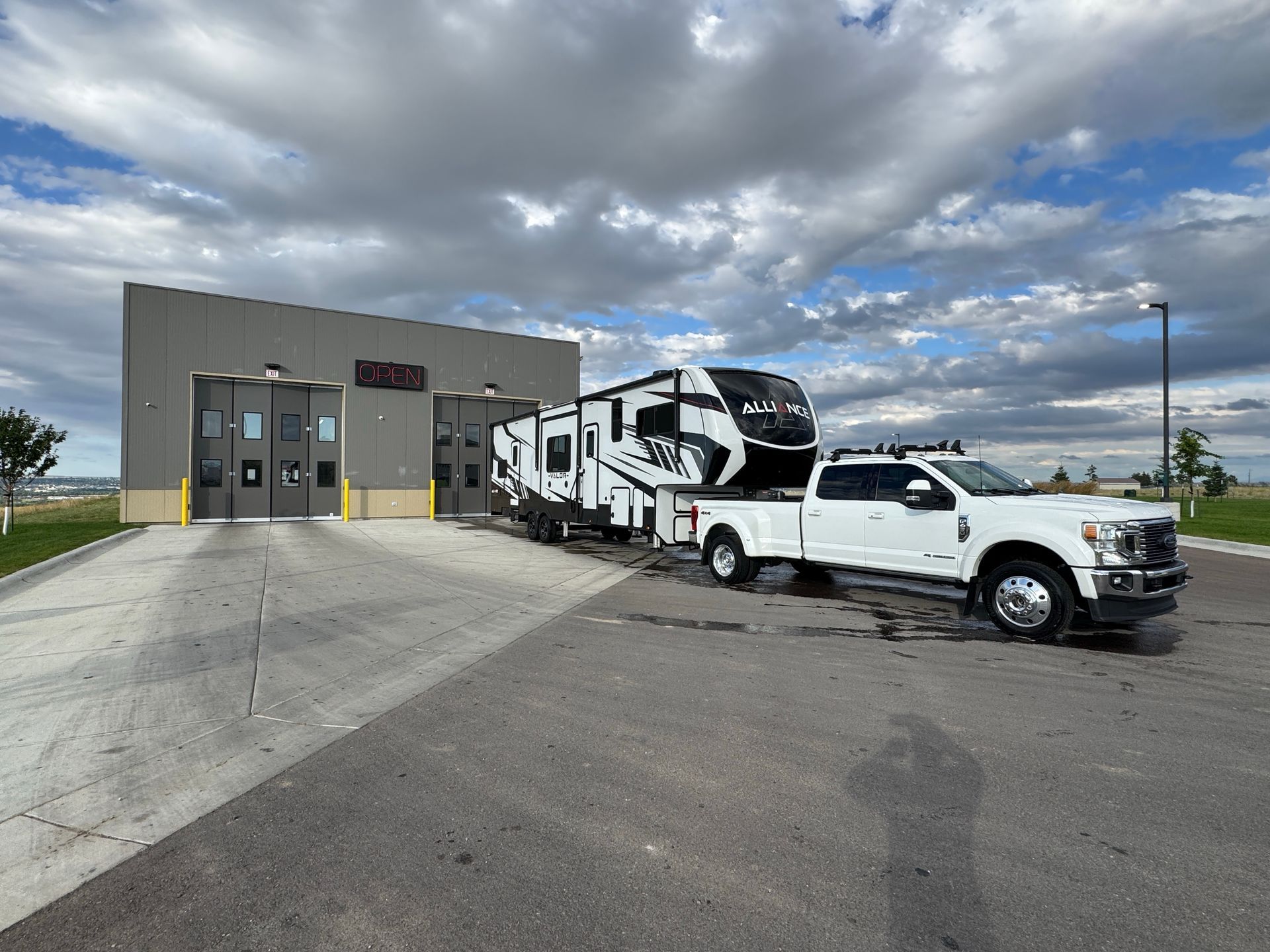 White pickup truck towing a black-and-white RV beside a gray building under cloudy skies.
