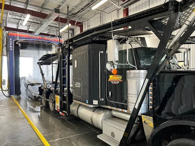 Black semi-truck in a service bay, connected to a wash system with hoses and equipment.