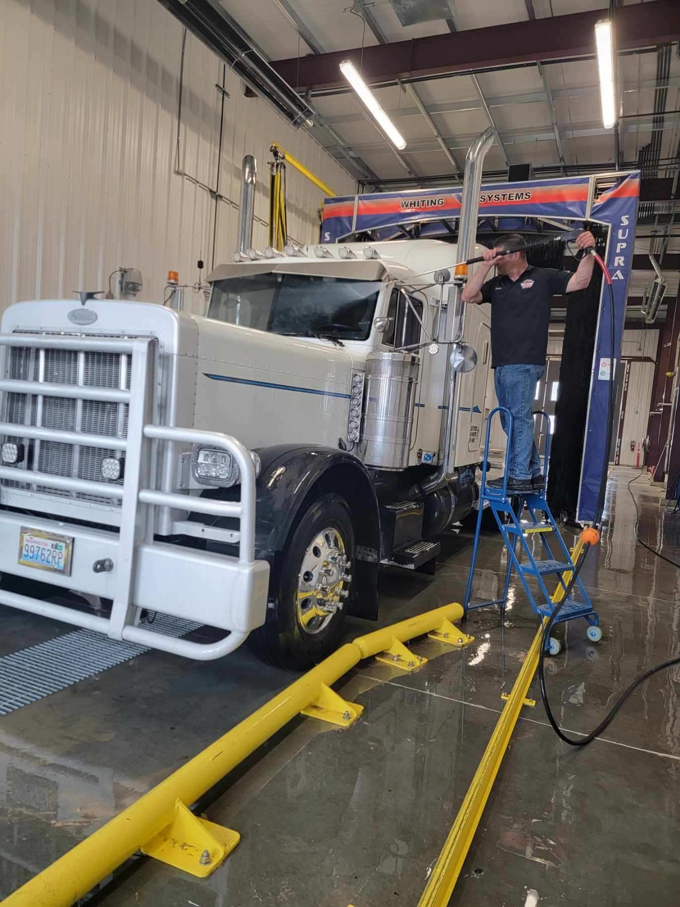 White semi-truck being washed in an industrial bay by two workers with cleaning equipment.