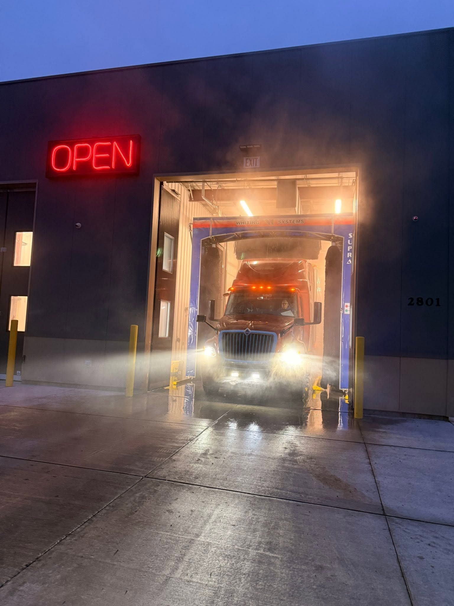 Truck entering a car wash at night beneath a red neon OPEN sign, water spraying on the wet floor
