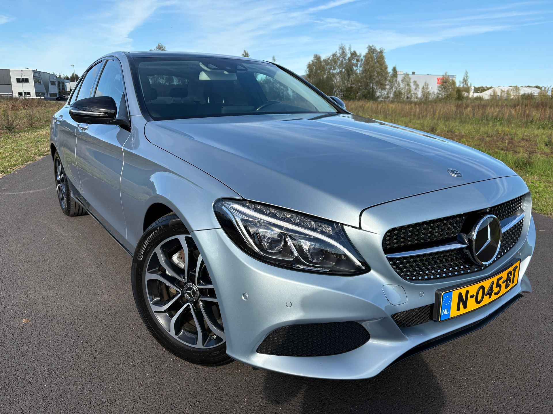 Een zilverkleurige Mercedes-Benz sedan staat geparkeerd in de buitenlucht op een zonnige weg.