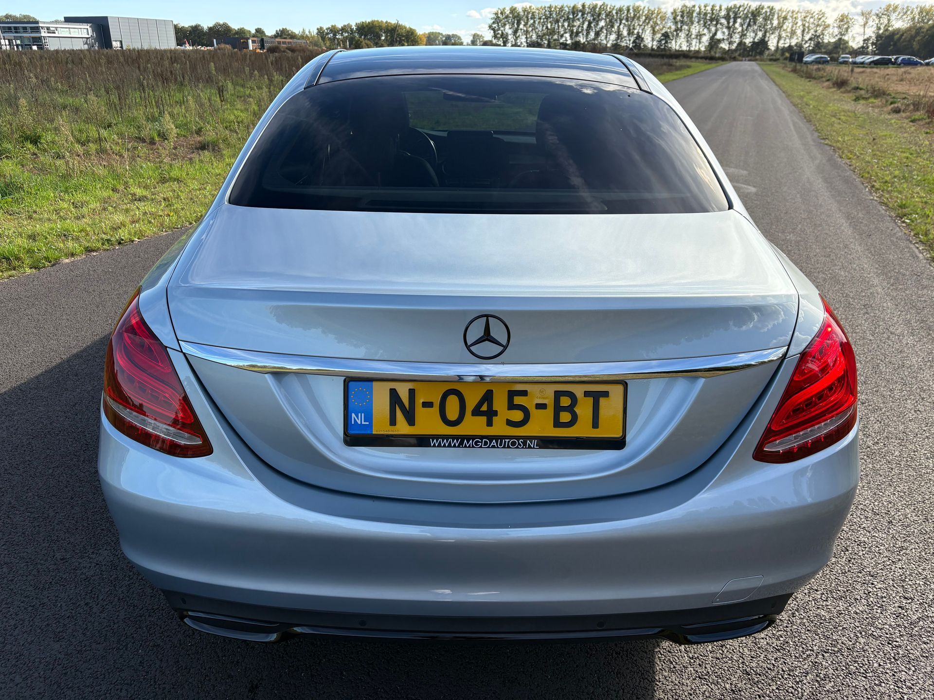 Achteraanzicht van een lichtblauwe Mercedes sedan, geparkeerd op een landweg, met een gele kentekenplaat.