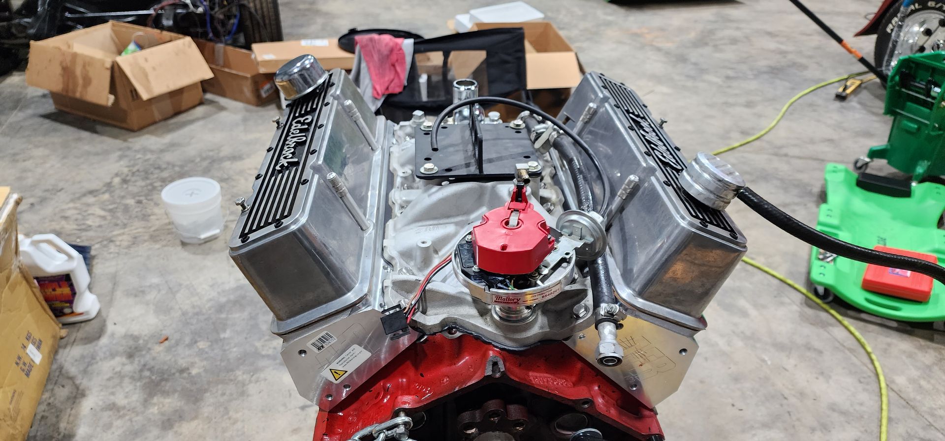 V8 engine on a workshop floor with chrome valve covers and a red intake manifold, surrounded by tools and boxes