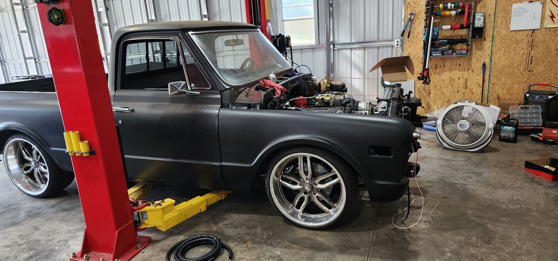 Black classic car in an auto shop beside a red lift, with tools and parts scattered around.