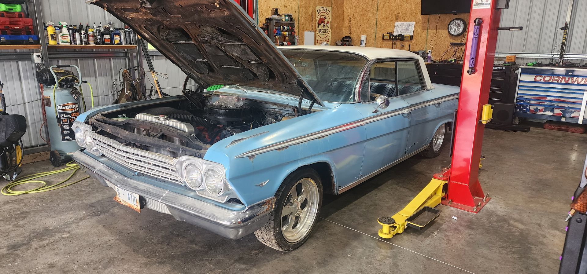 Blue classic car in a garage, hood open on a lift, with tools and shop equipment nearby.