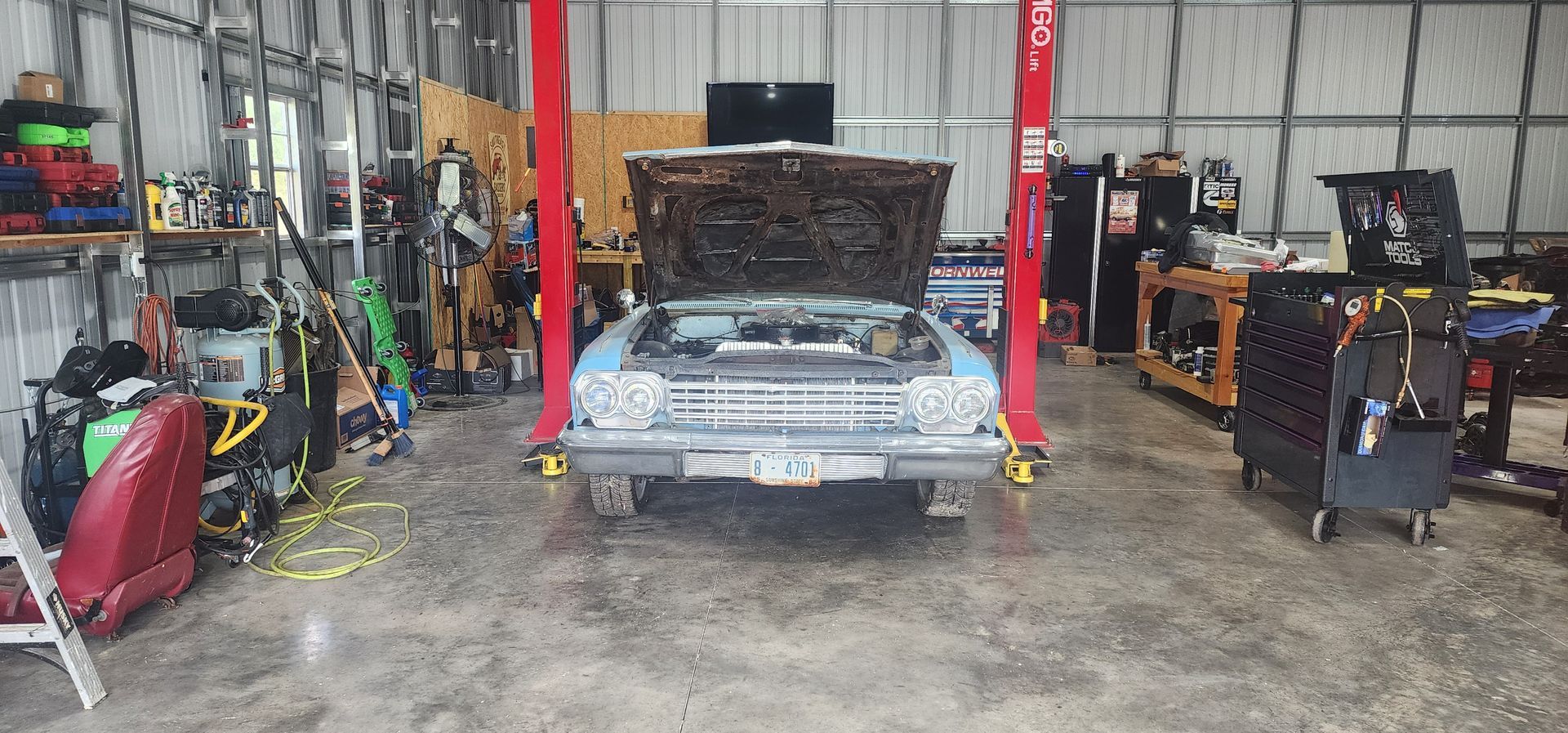 Classic car in a garage with hood open, surrounded by tools and equipment.