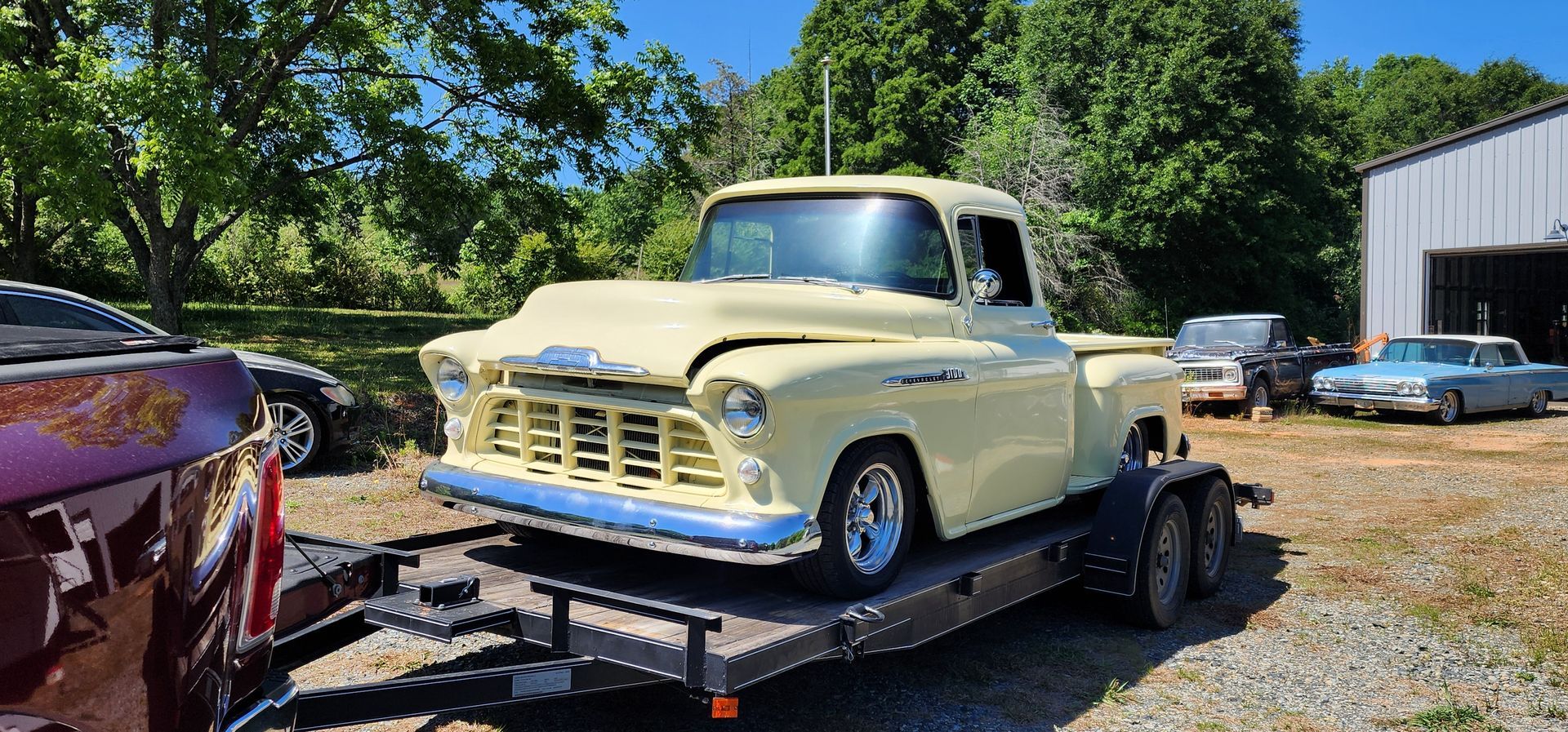 Vintage cream pickup truck on a flatbed trailer at an outdoor car lot, with other classic cars nearby