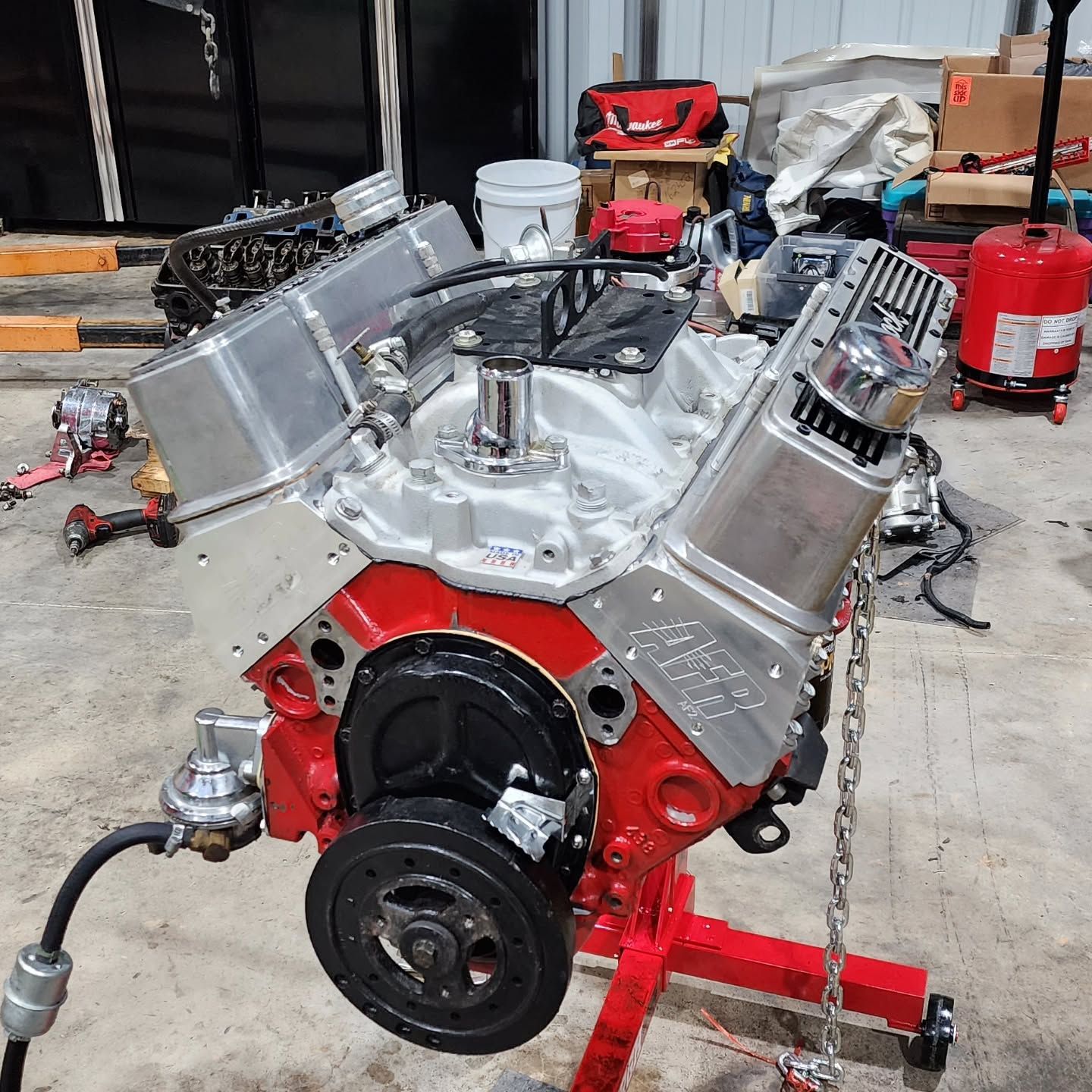 Polished V8 engine on a red stand in a workshop, with silver valve covers and black front pulley.