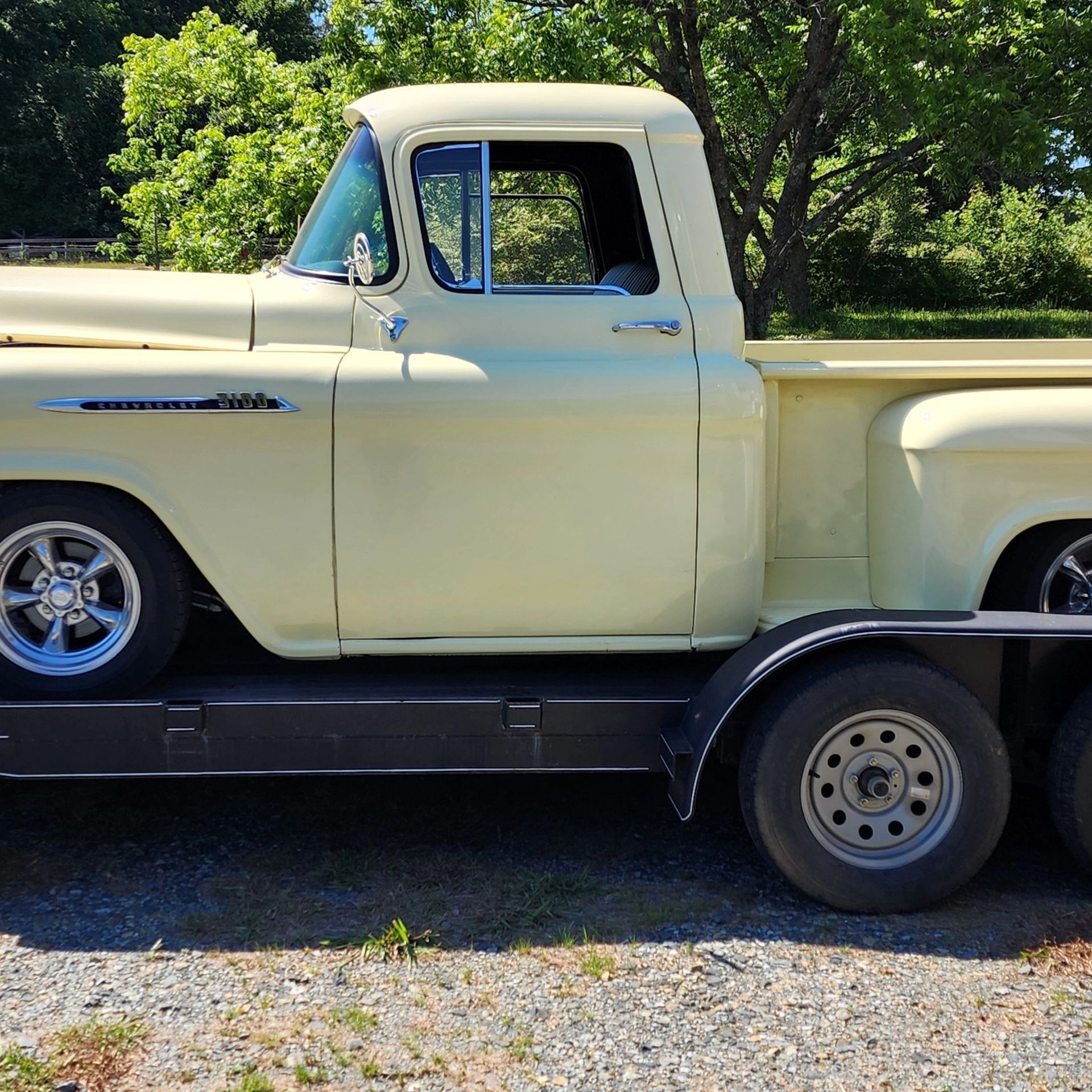 Vintage cream pickup truck on a flatbed trailer, parked outdoors among green trees.