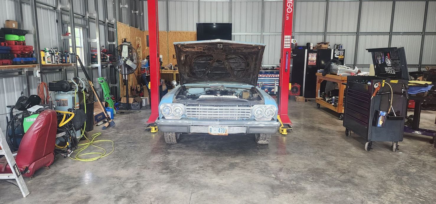 Classic car in an auto repair shop with its hood open, surrounded by tools and equipment.