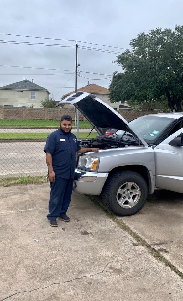 Mechanic in blue uniform next to silver pickup with open hood, cloudy day.