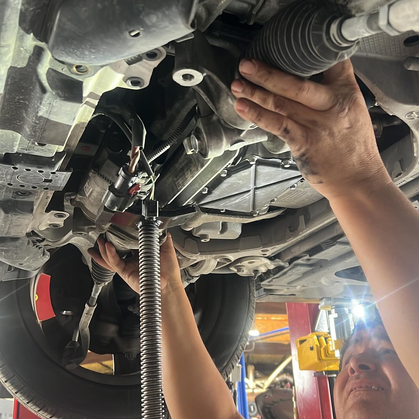 Hands working on car's underside, likely auto repair. Tools visible. Bright overhead light. | William’s Auto Repair