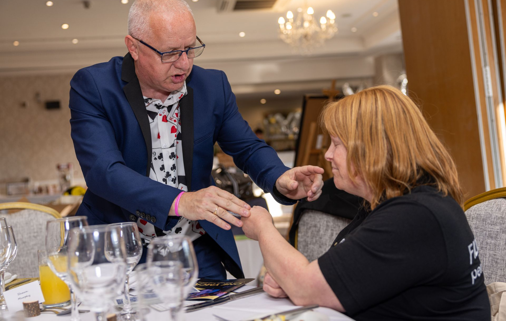 A man in a blazer leans over a table to examine a ring on a person's hand in an indoor dining setting.