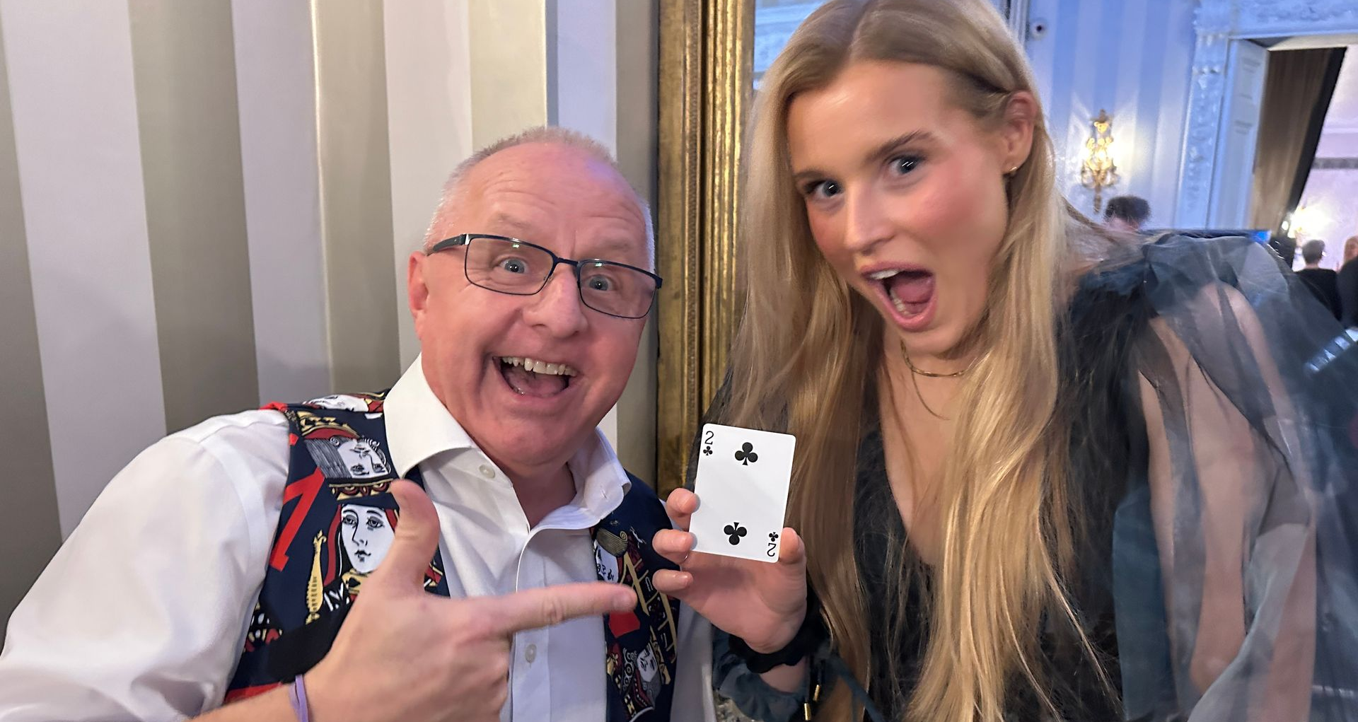 A man pointing at a playing card held by a woman, both with enthusiastic expressions, in an indoor room.