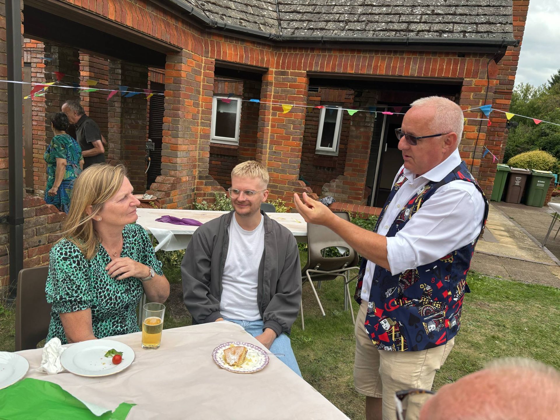 A group of people sit and stand around a garden table, listening to a man gesturing while holding a small object.