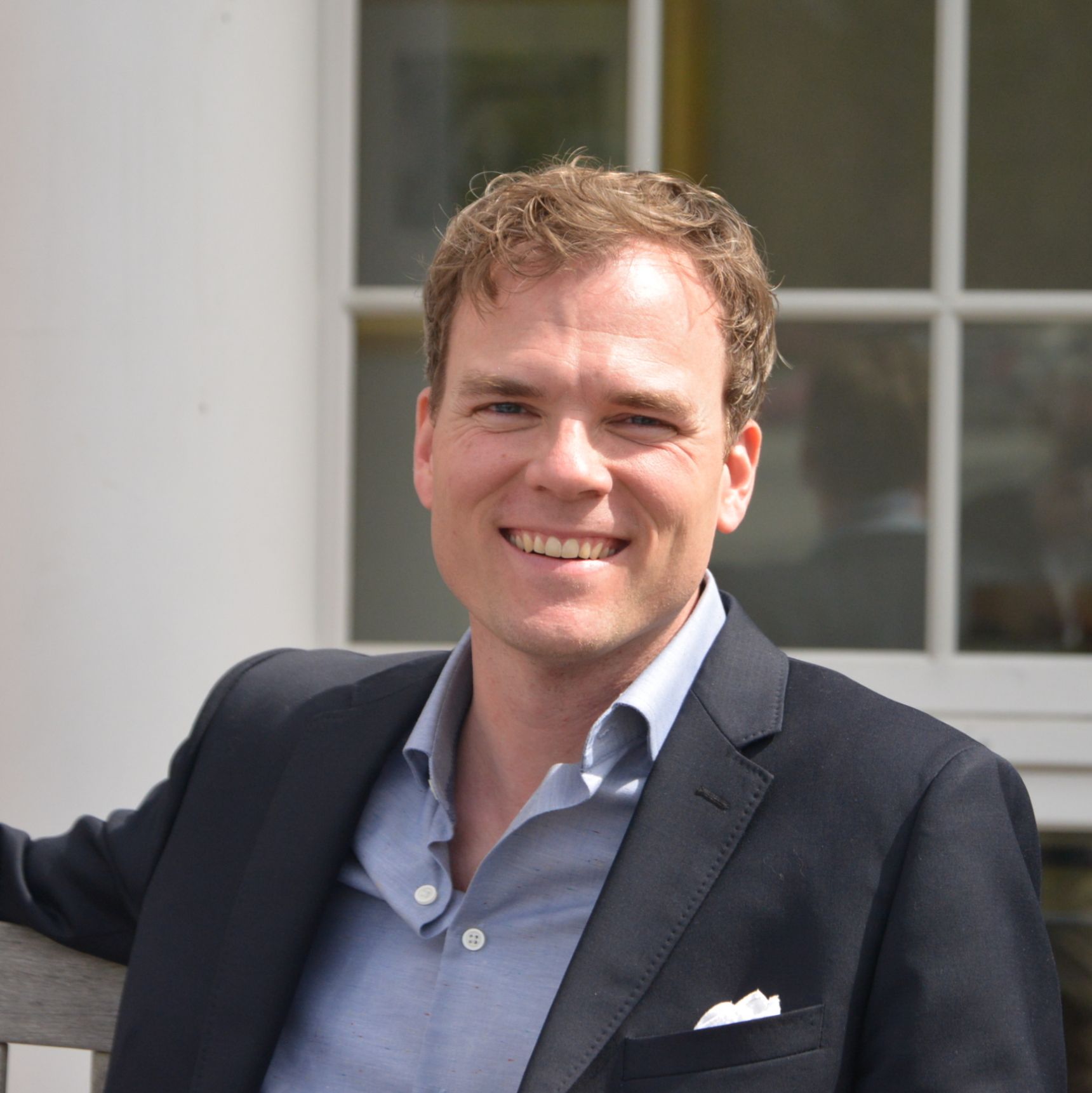 Man in suit jacket smiles at the camera against a building