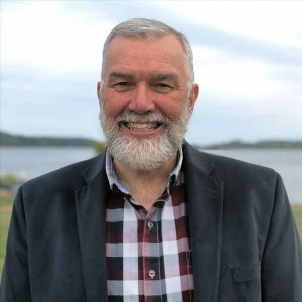 Man with gray beard smiles, wearing checked shirt and blue blazer, outdoors near water.