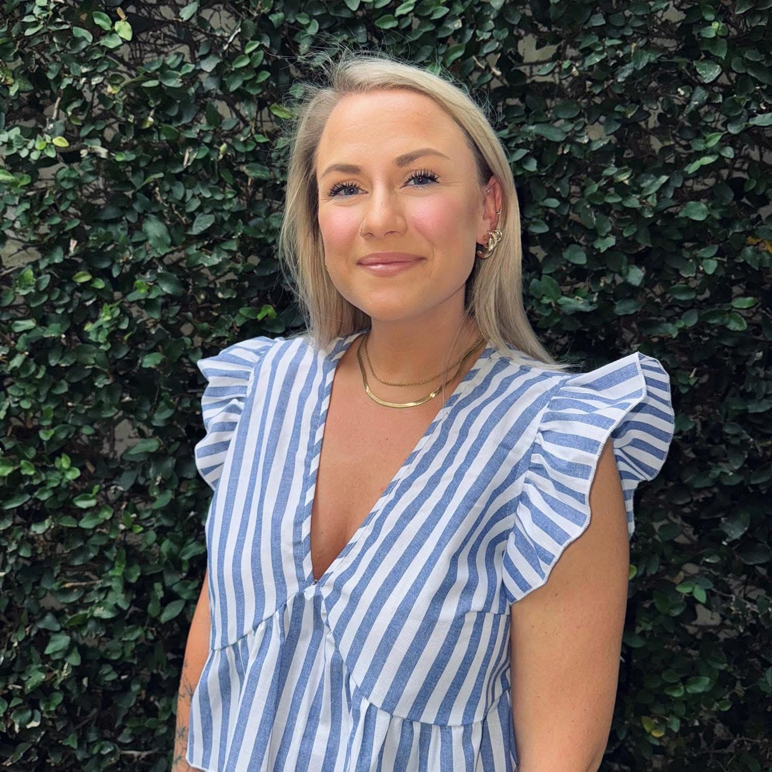 Woman with blonde hair smiles, wearing a blue and white striped shirt, outdoors near green foliage.